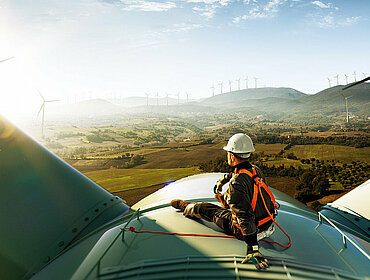 Ein glücklicher Ingenieur, der sich nach gelungener Arbeit über seinen Erfolg freut. Er steht oben auf einer Windkraftanlage und blickt auf eine schöne Sonnenuntergangslandschaft.