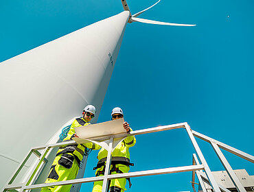 Zwei Arbeiter in leuchtender Sicherheitskleidung überprüfen Pläne in der Nähe eines hohen Windkraftwerks an einem sonnigen Tag.